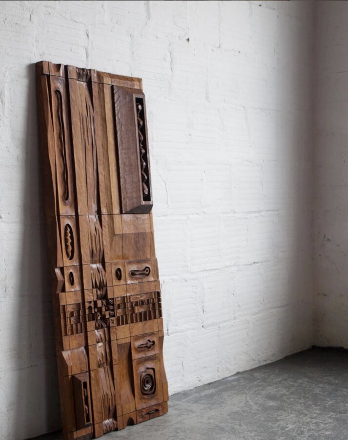 A carved wood panel by Leroy Setziol leaning against a whitewashed brick wall.